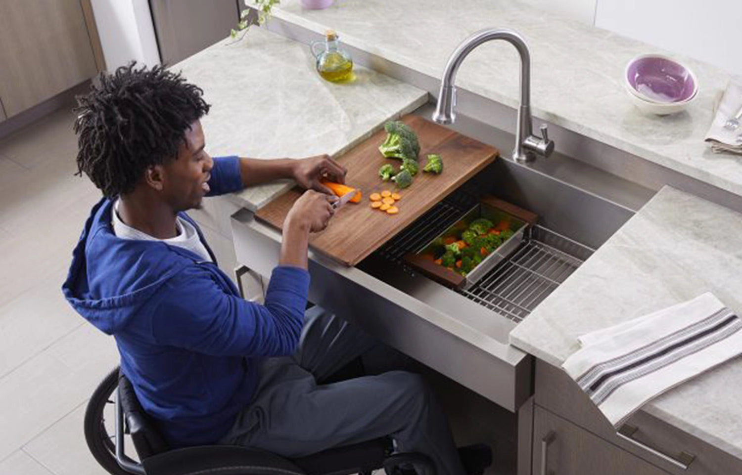 A boy in a wheelchair is slicing vegetables on a wooden cutting board, utilizing an ADA-compliant kitchen sink.